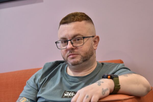 A photograph of Paul sat on a orange couch in a light purple room