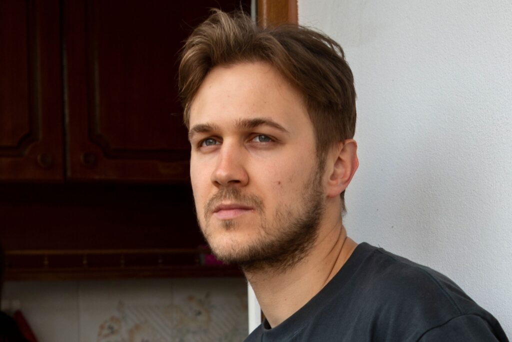 photograph of a man standing in a doorway, looking towards the camera. Behind him stands a woman in a kitchen with her back to us.