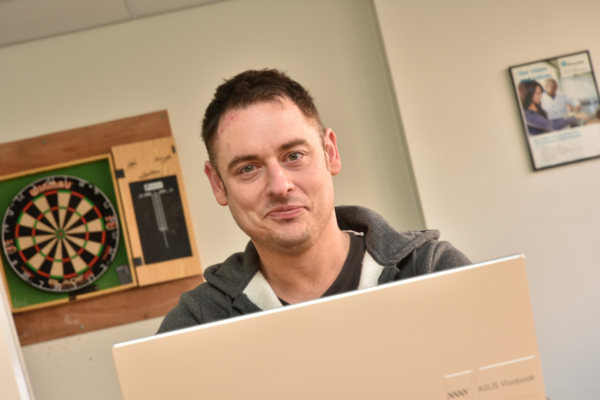 Photograph of a man sat in front of a laptop looking towards the camera