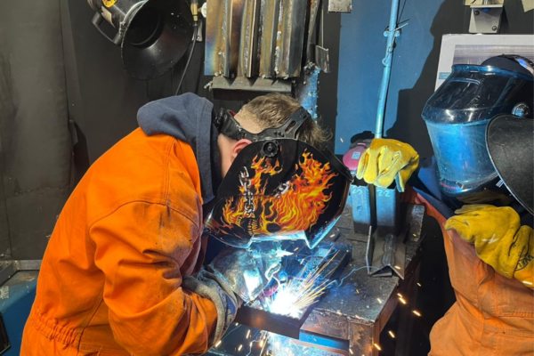 Photograph of a person wearing an orange jump suit and a welding mask welding a piece of metal