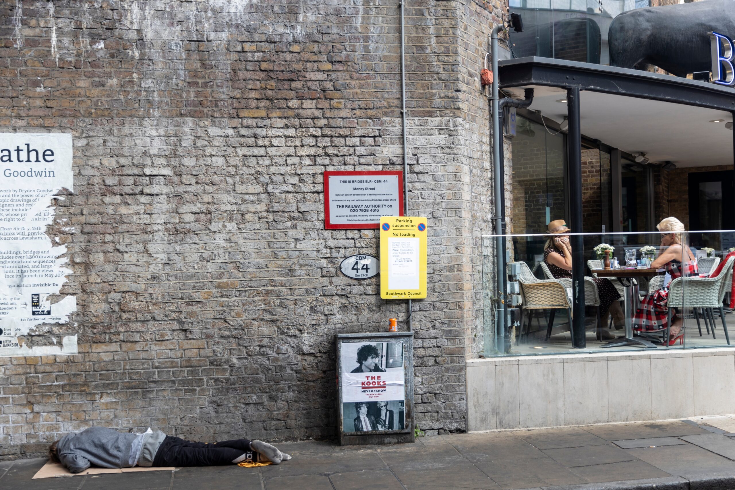 World Homeless Day blog post image of a homeless man laying on the ground sleeping as you can see two woman drinking and drinking happily on a balcony 