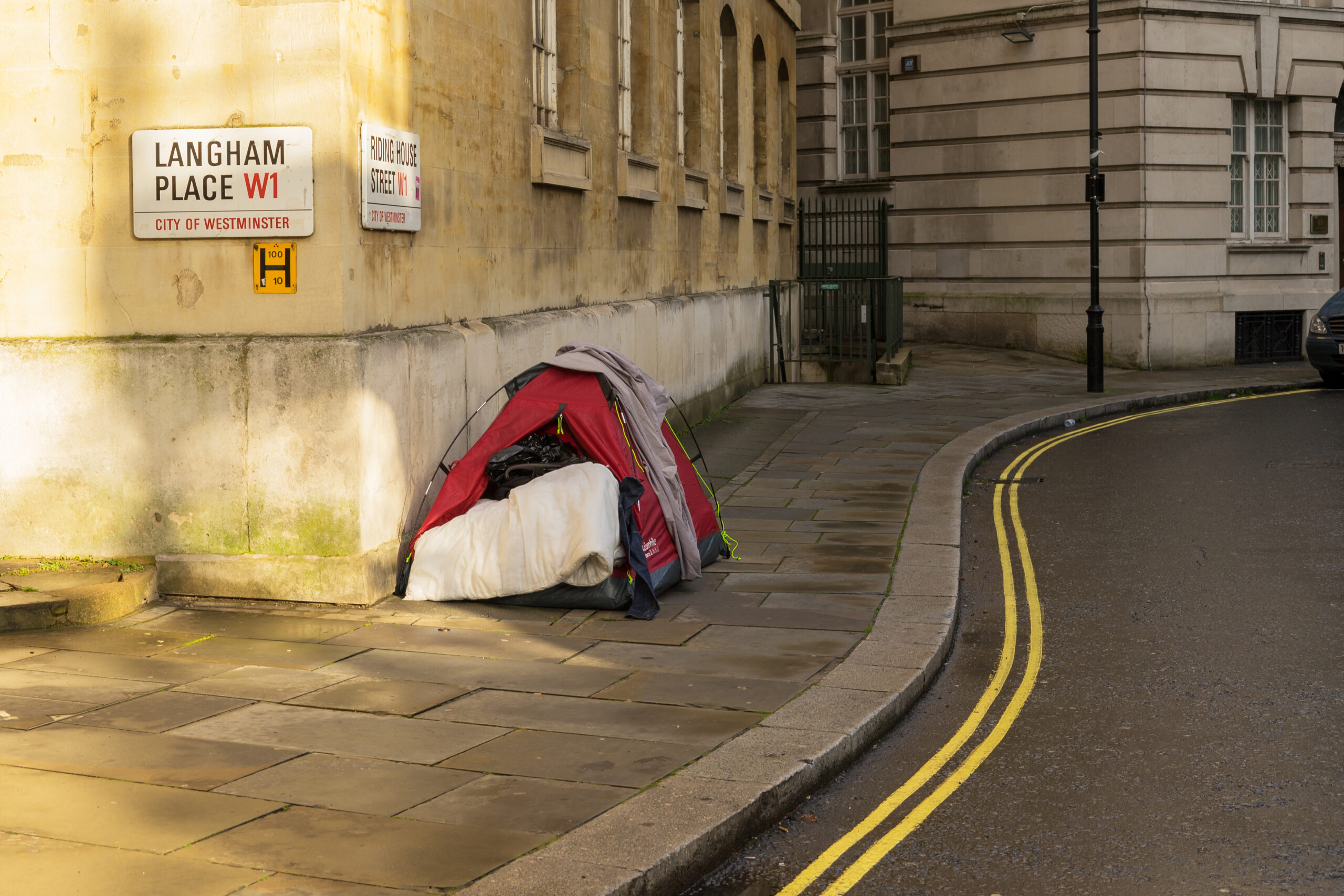 Photograph of a london street with a dishevelled tent on it- implying someone who is homeless is living in the tent.  